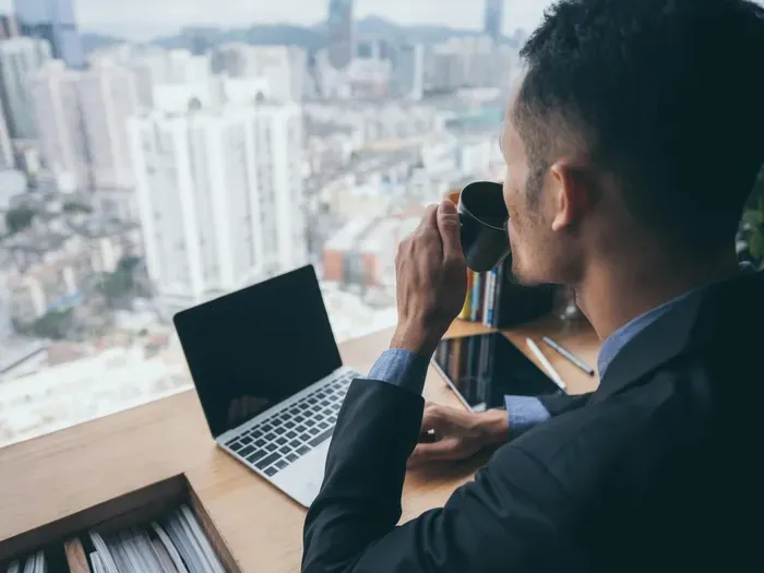 Entrepreneur in a suit drinks coffee at a desk with laptop overlooking skyline; City Service List supports business
